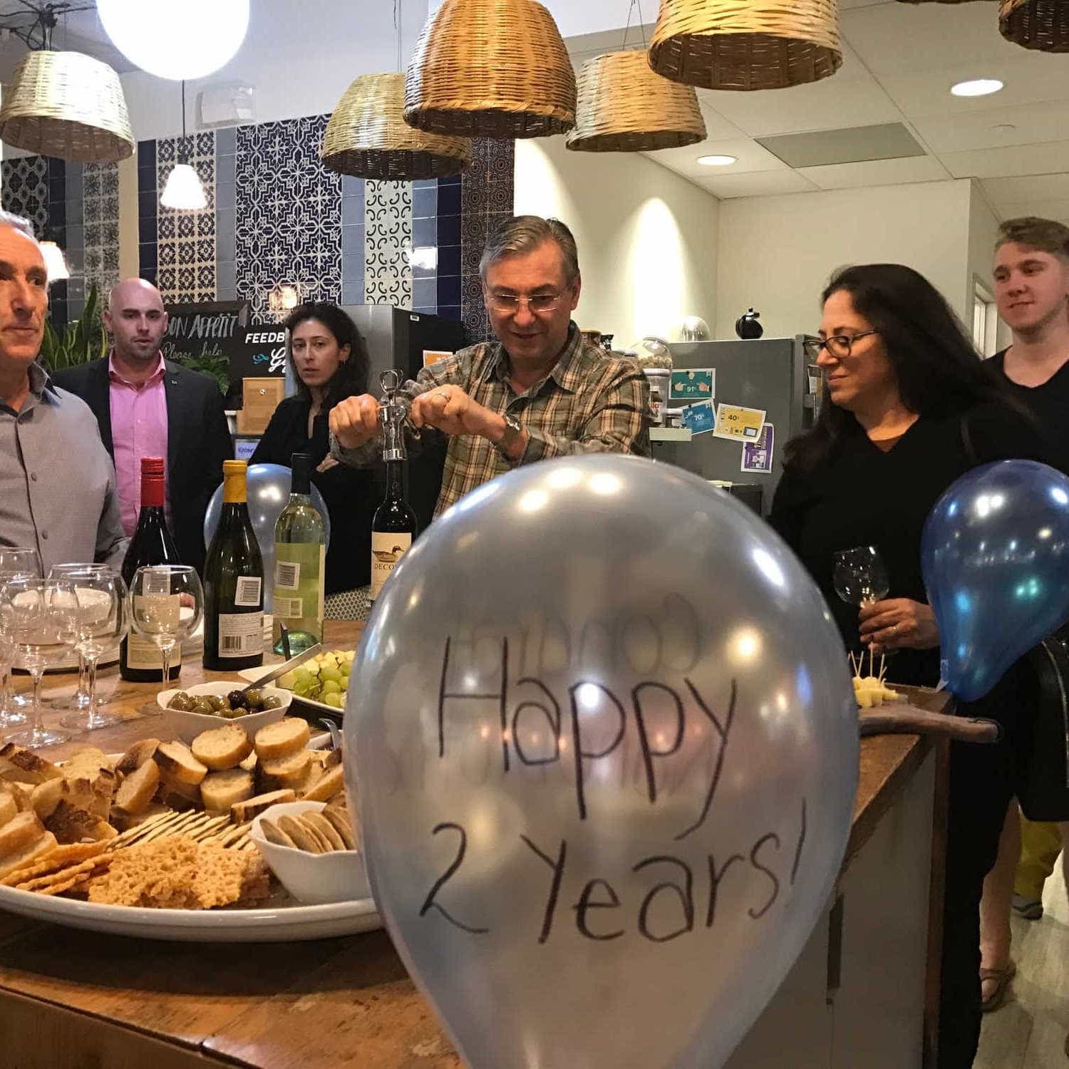 Many members celebrating a 2 year anniversary in NGIN Workplace's kitchen and dining area, with food, wine, and balloons.